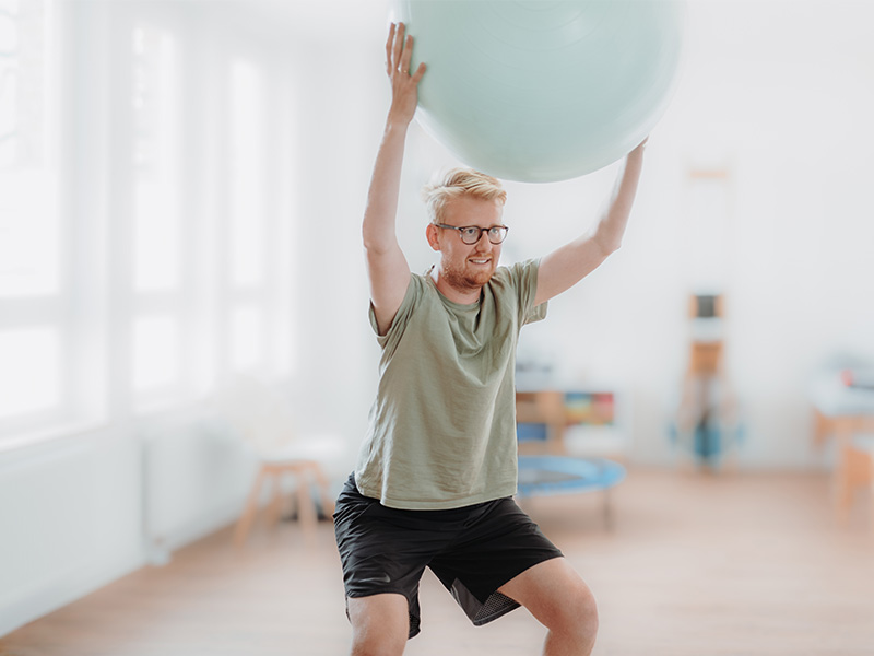 Patient mit Gymnastikball über dem Kopf haltend im Rahmen der Krankengymnastik